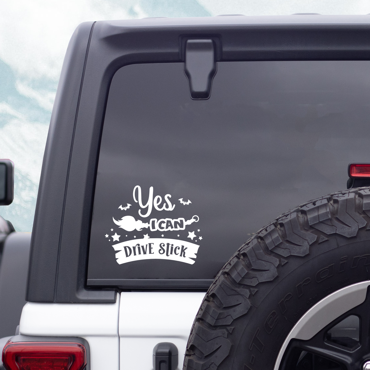 A white vinyl decal with the text 'Yes I CAN Drive Stick' and a small witch hat illustration, applied to the rear window of a white vehicle.