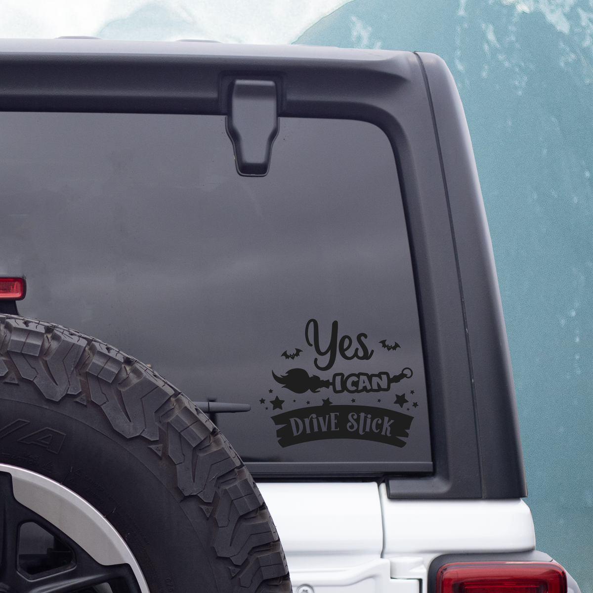 A black vinyl decal with the text 'Yes I CAN Drive Stick' and a witch hat graphic, applied to the rear window of a dark SUV, with a spare tire visible on the left.