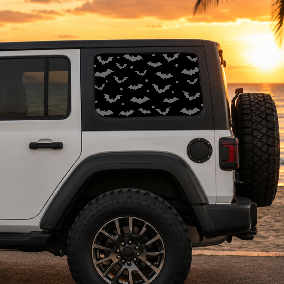 A white Jeep parked on a beach at sunset, with a black window shade featuring a repeating bat pattern installed on the rear side window.