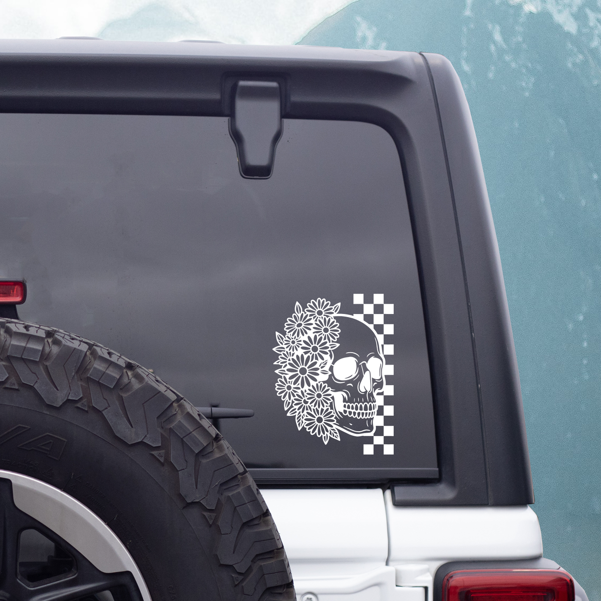 A white vinyl decal featuring a skull decorated with a floral wreath and a black-and-white checkered square, shown applied to the rear window of a vehicle.