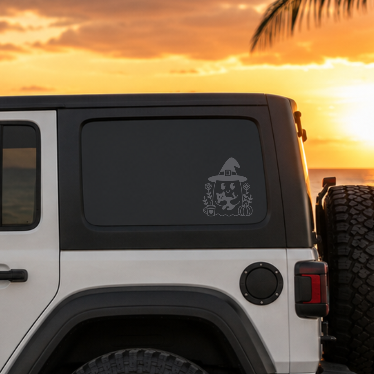 A white Jeep Wrangler parked outdoors at sunset, with a gray decorative decal of a witch and pumpkin on the rear side window. The background shows a warm orange sky and ocean at sunset.