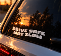 A white vinyl bumper sticker with the text 'DRIVE SAFE NOT SLOW' applied to the rear window of a silver car, reflecting a warm sunset sky and trees.