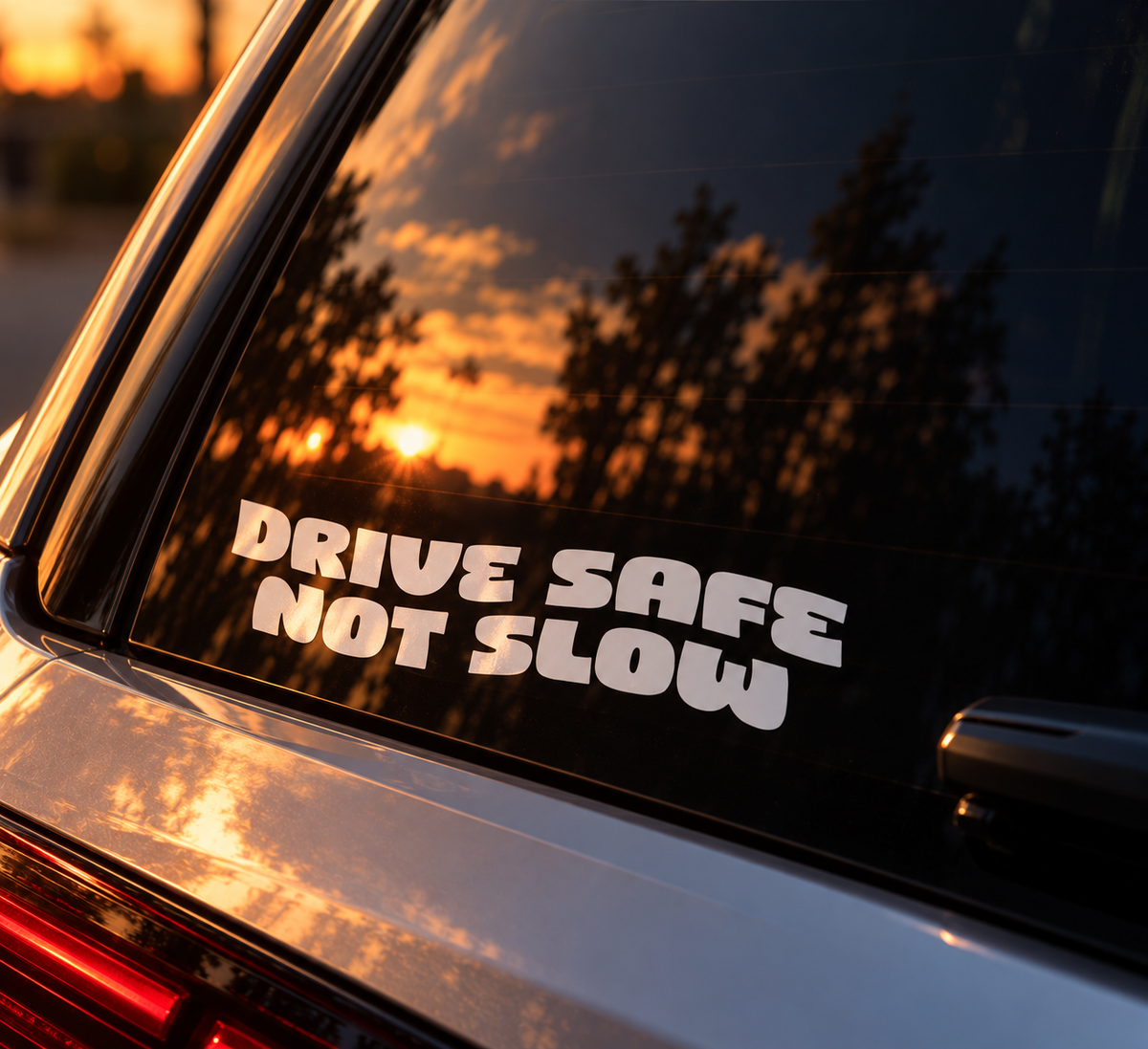 A white vinyl bumper sticker with the text 'DRIVE SAFE NOT SLOW' applied to the rear window of a silver car, reflecting a warm sunset sky and trees.