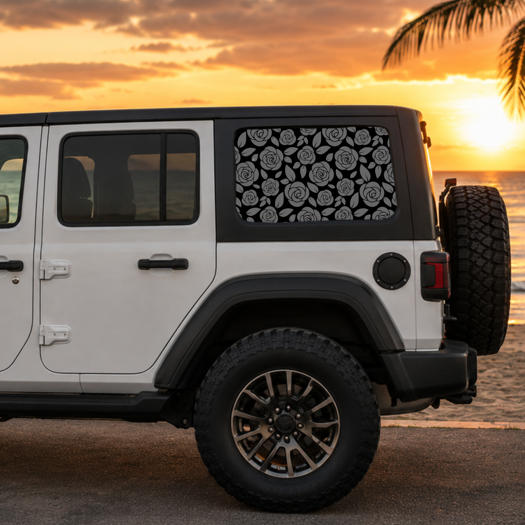 A white Jeep Wrangler parked outdoors at sunset near the beach, showing a black-and-white floral patterned sun shade installed on the rear side window.