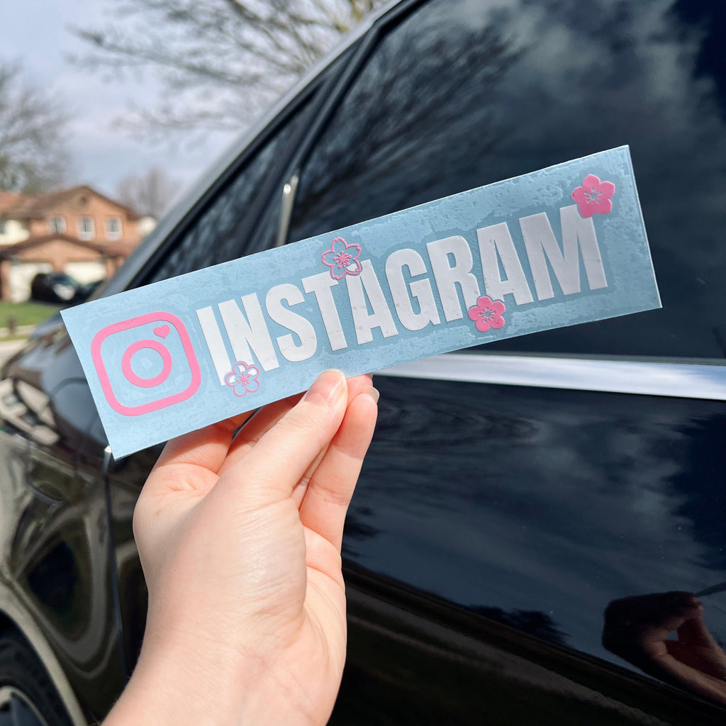 Hand holding a 'INSTAGRAM' sticker with pink flowers against a car window.